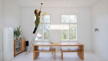 A long shot of a woman standing on a ladder in her minimalist kitchen installing the kupu smoke alarm. 